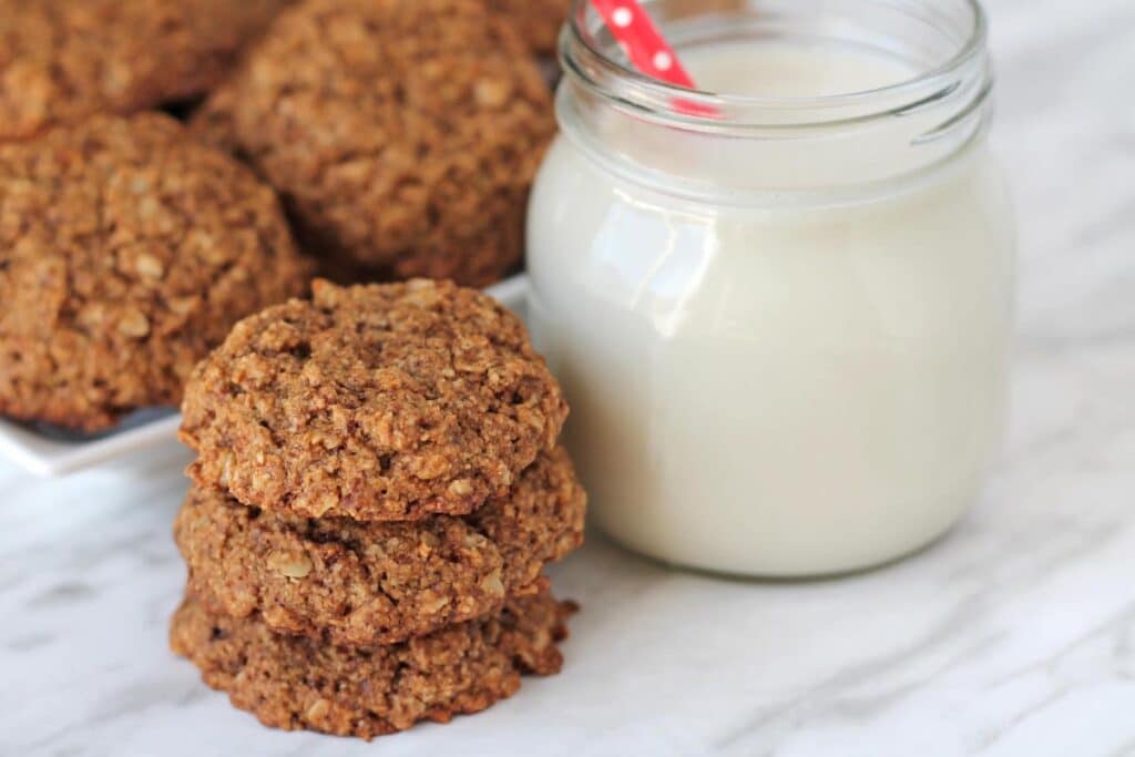 Three oatmeal cookies stacked on top of each other, more cookies sit behind the stack and a glass of milk sits to the right.