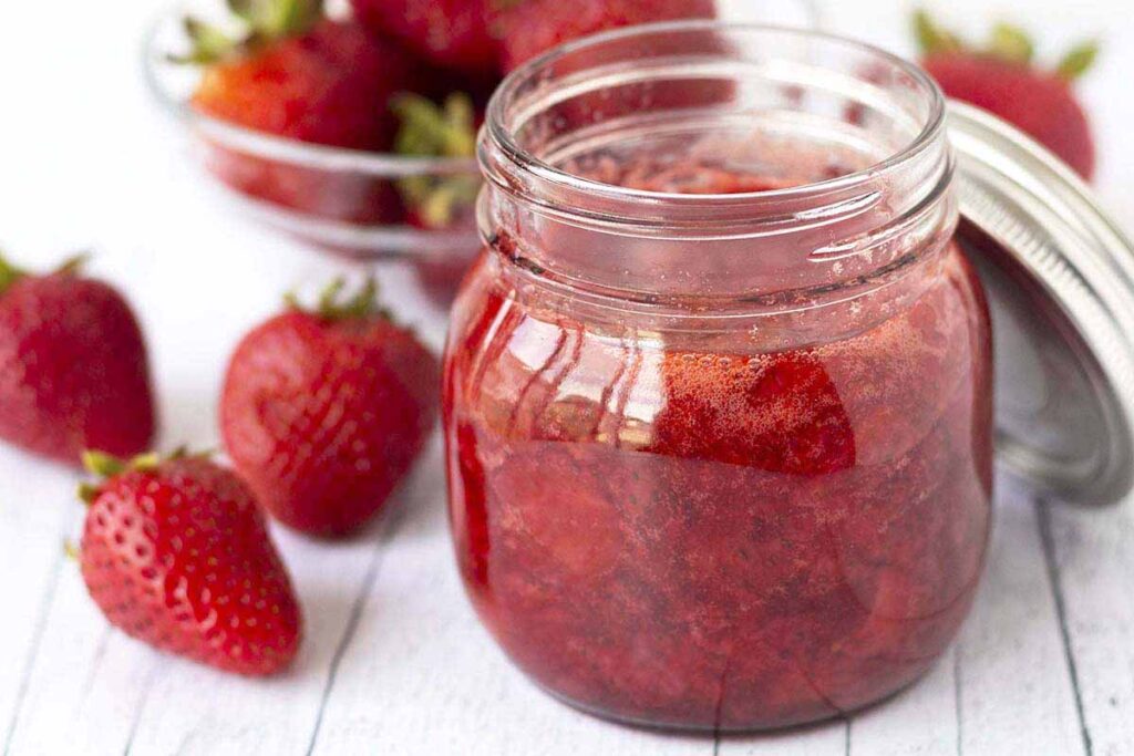 A jar of fresh strawberry sauce sitting on a wood surface, fresh strawberries sit to the left of the jar.
