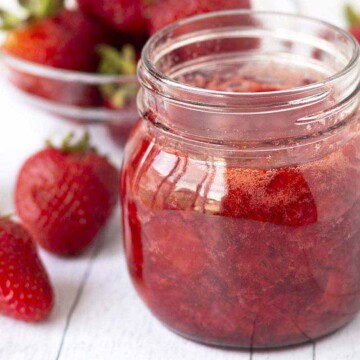 A jar of fresh strawberry sauce sitting on a wood surface, fresh strawberries sit to the left of the jar.