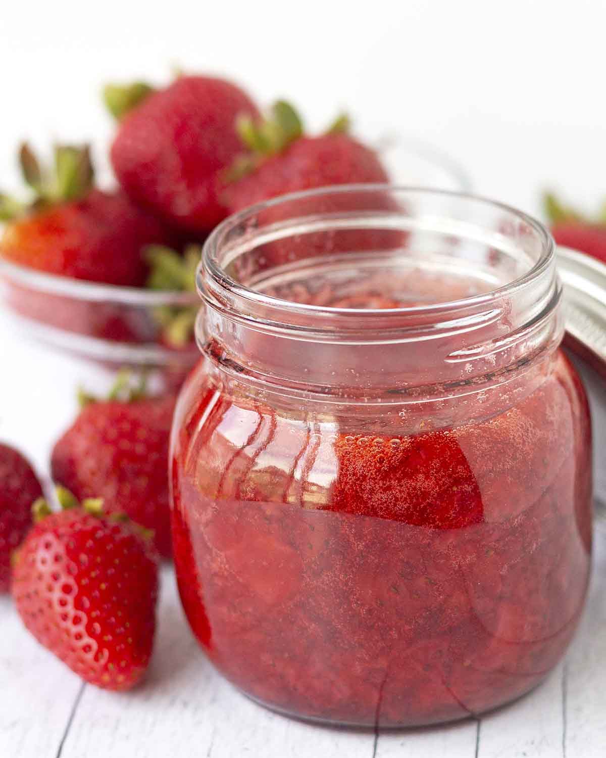 A glass jar of freshly made homemade strawberry sauce in a glass jar, there are fresh strawberries around the jar.