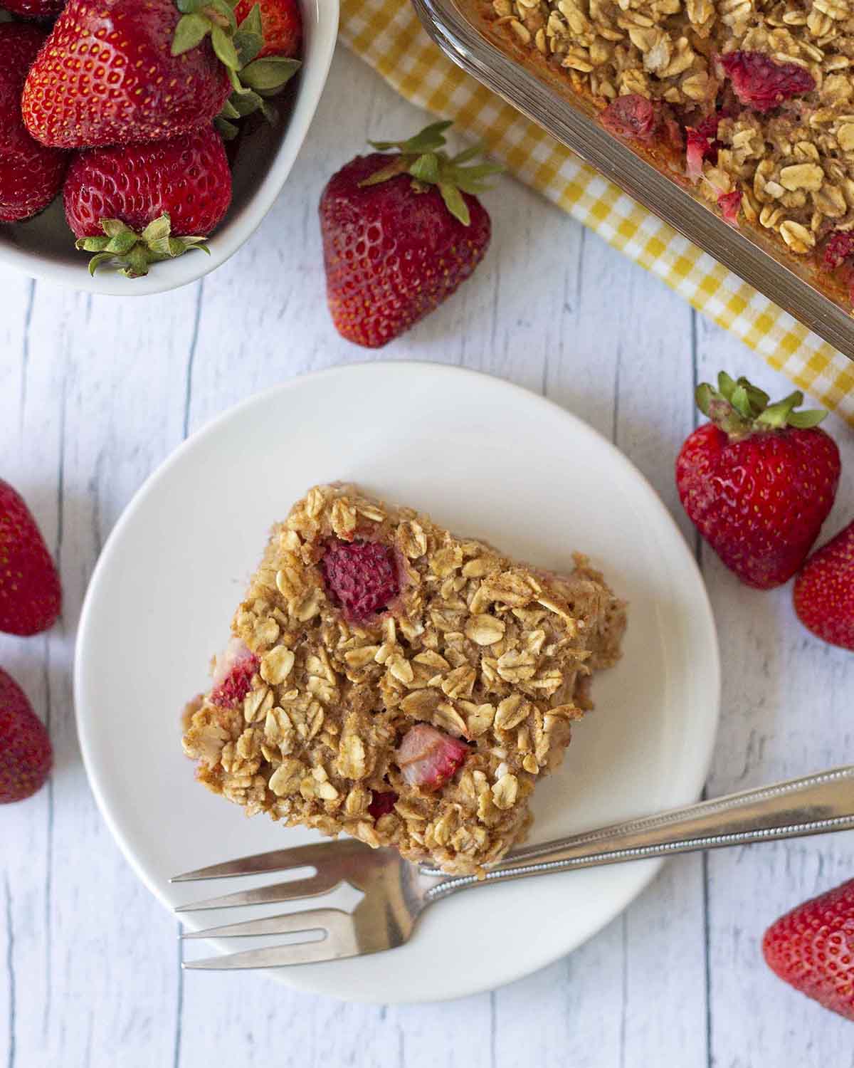 An overhead shot showing a slice of strawberry baked oatmeal on a plate, fresh strawberries are around the plate.