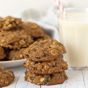 A stack of three oatmeal raisin cookies, more cookies are on a plate behind the stack, a glass of milk sits to the right.