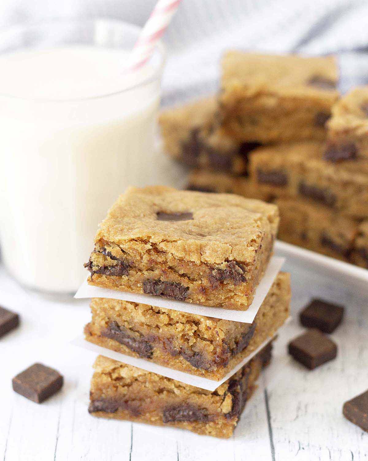 A stack of three gluten-free blondies, a glass of milk with a pink straw in it and more blondies sit behind the stack.