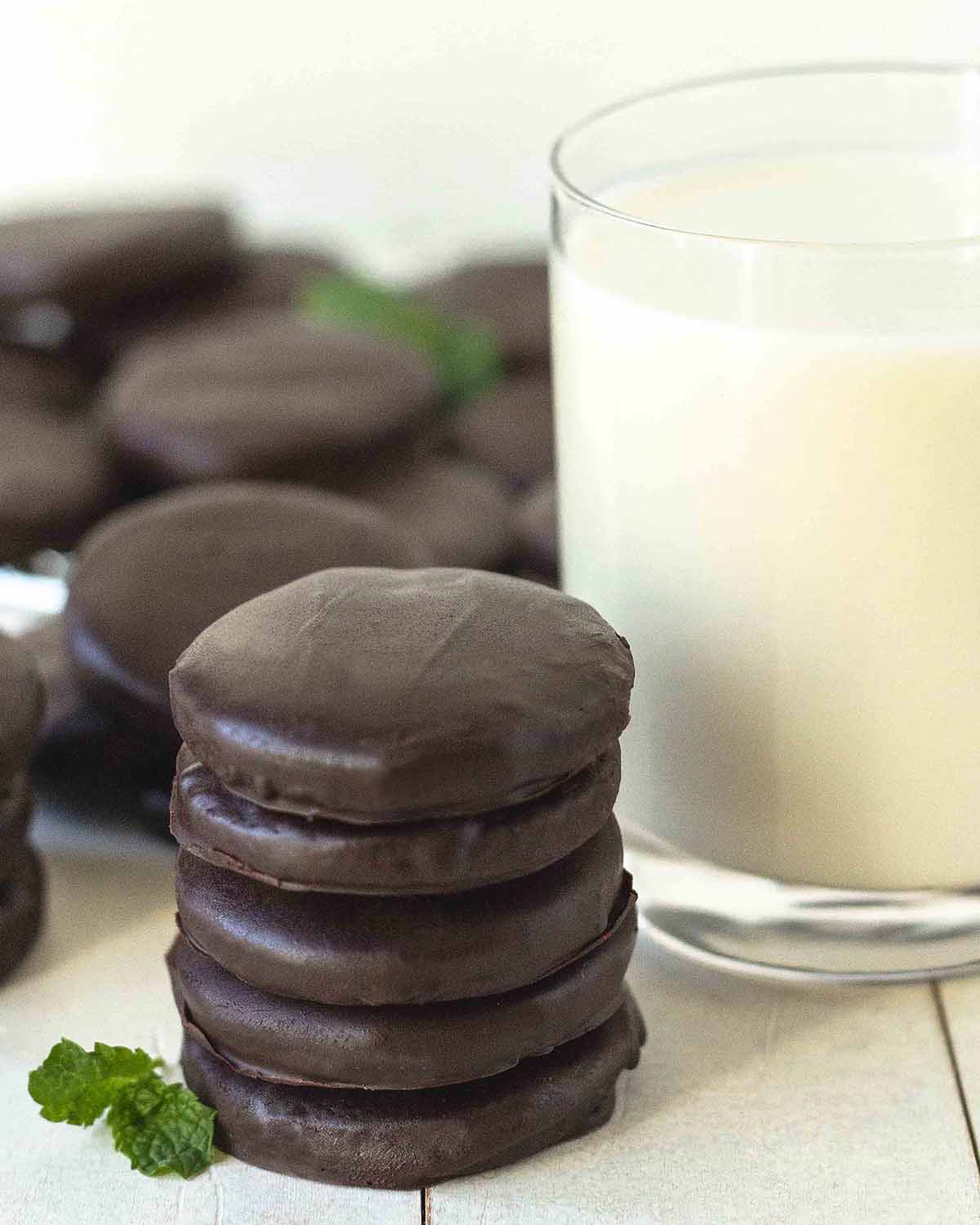 A stack of 5 chocolate mint dipped cookies sitting on a white table. More cookies and a glass of milk sit behind the cookies.