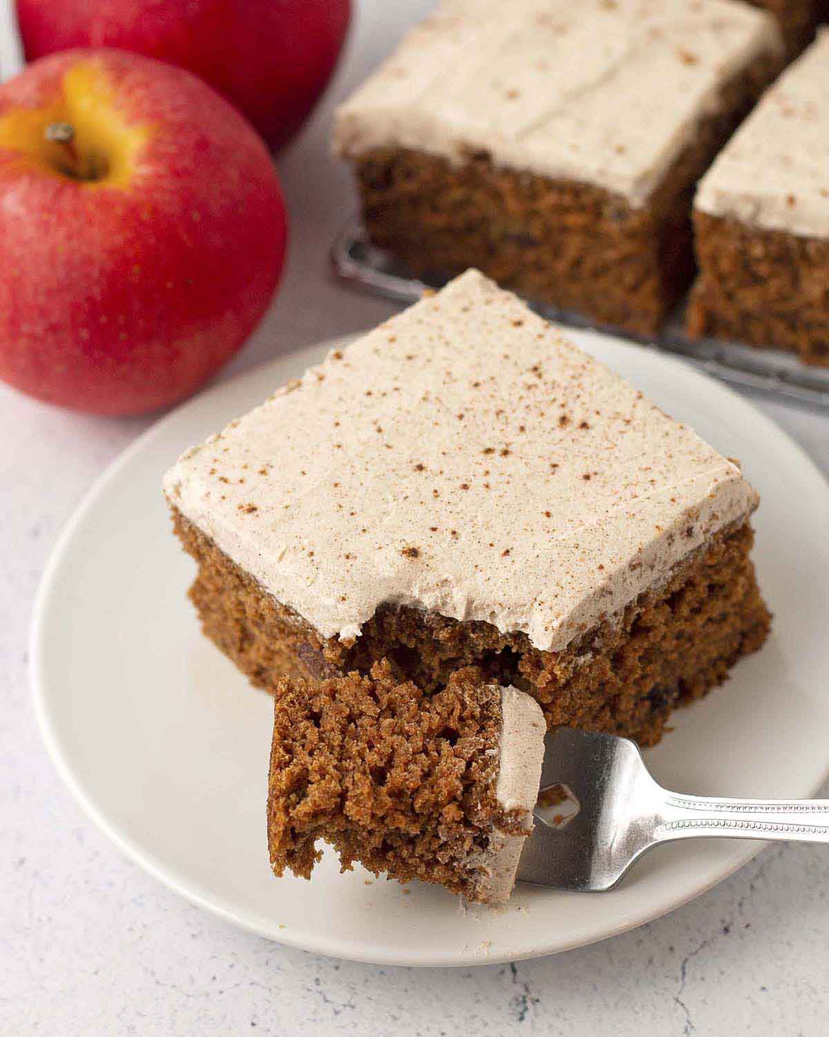 A square of eggless applesauce cake on a white plate, a piece of cake is on a fork that is on the plate in front of the cake.