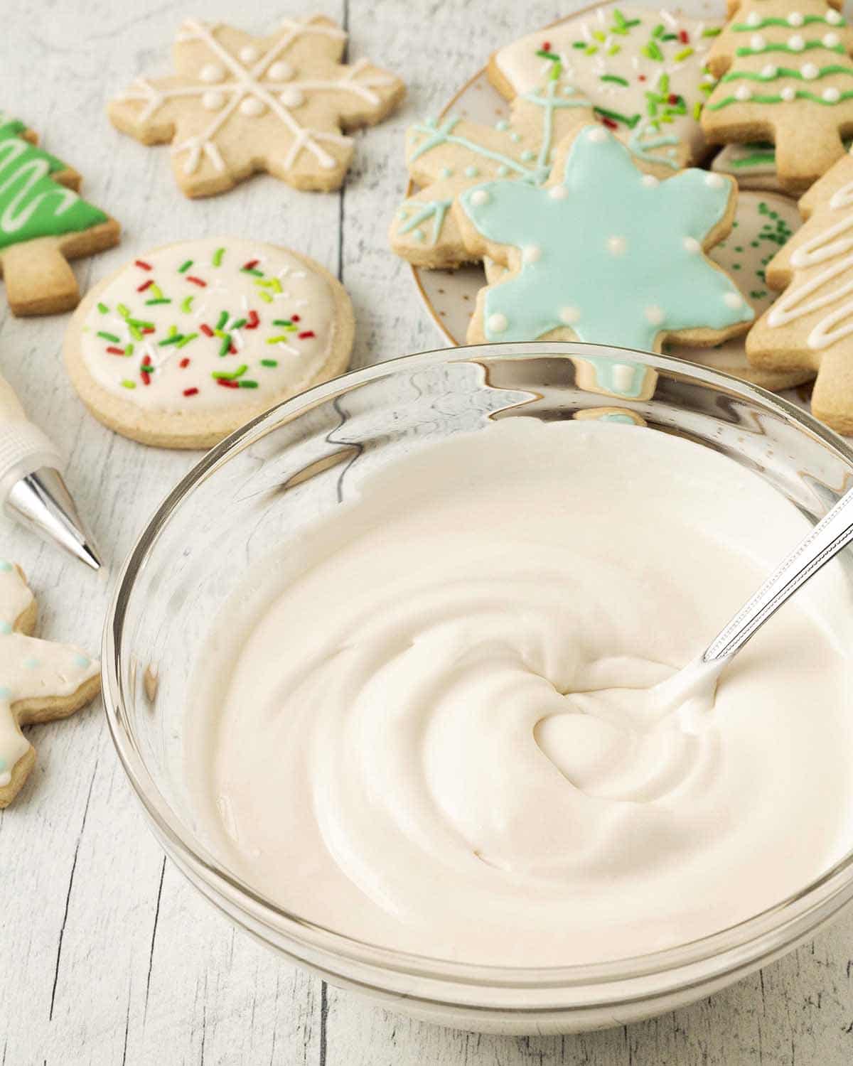 A bowl of vegan gluten-free royal icing on a white table, decorated cookies sit behind the bowl.