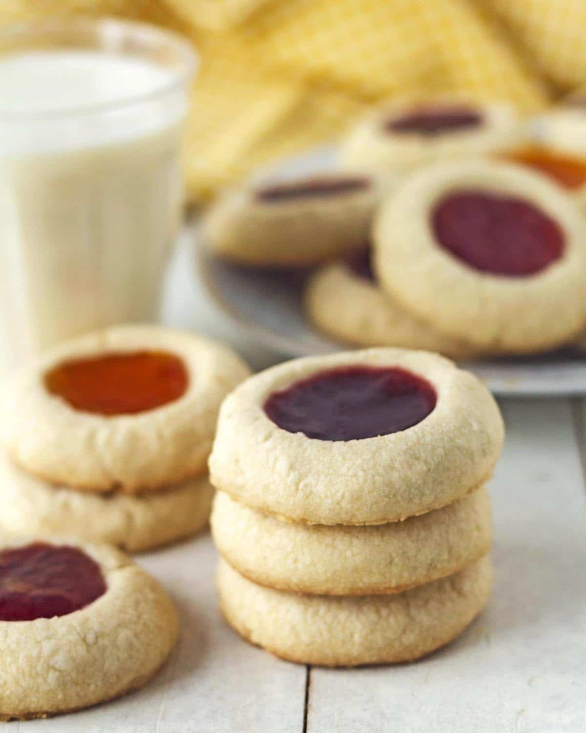 Three vegan gluten-free thumbprint cookies stacked on each other, more cookies sit on a plate behind and beside the stack.