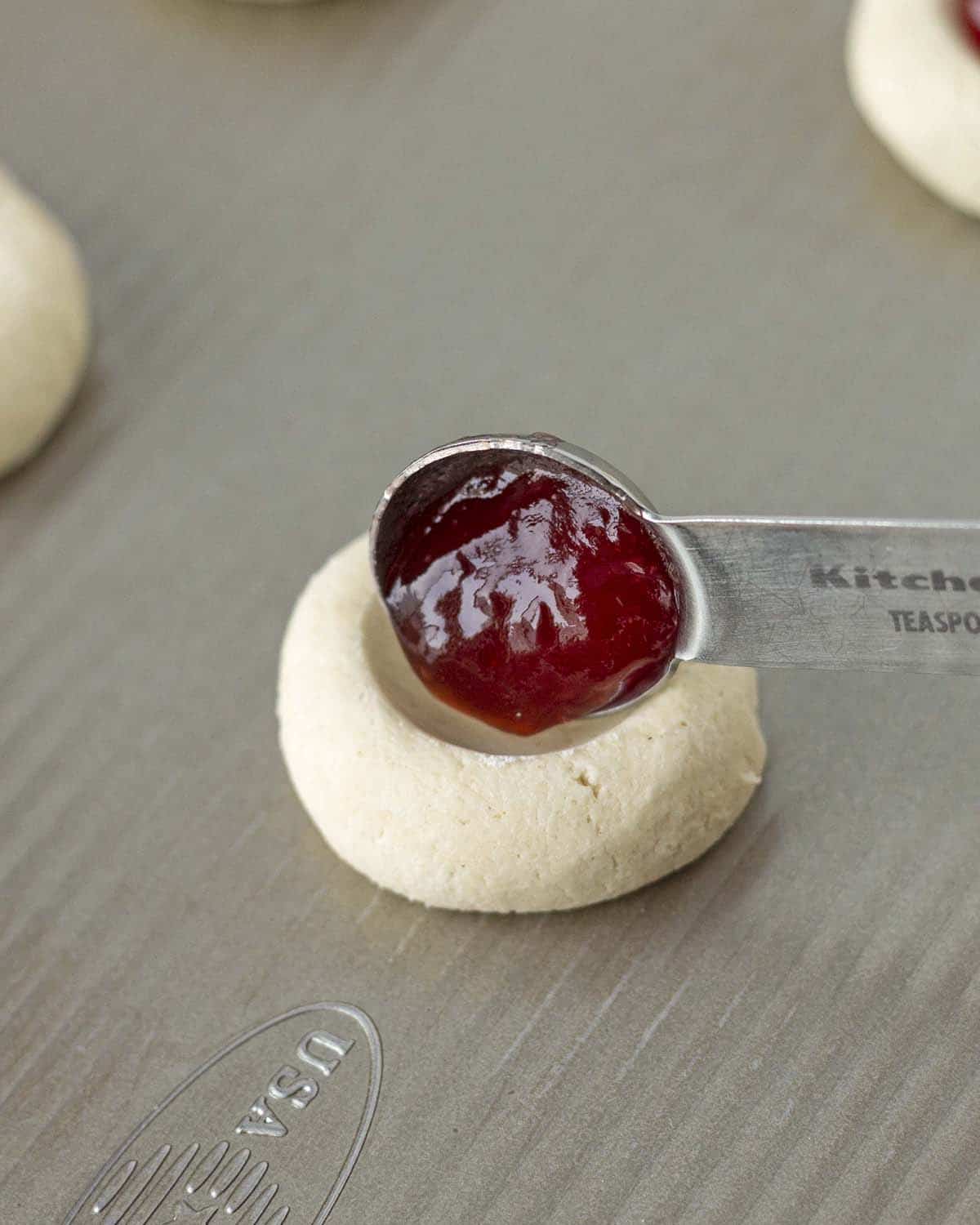 A measuring spoon putting raspberry jam into a thumbprint cookie before it bakes.