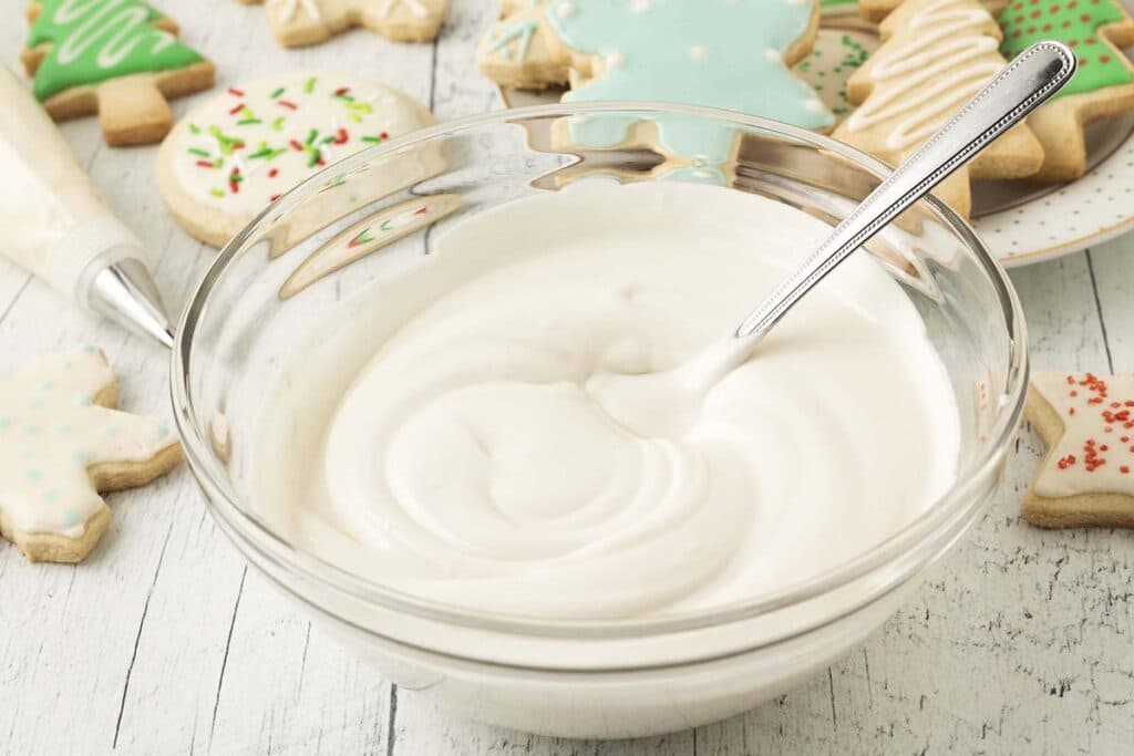 A glass bowl of vegan royal icing with a spoon in it, decorated cookies sit behind the bowl.
