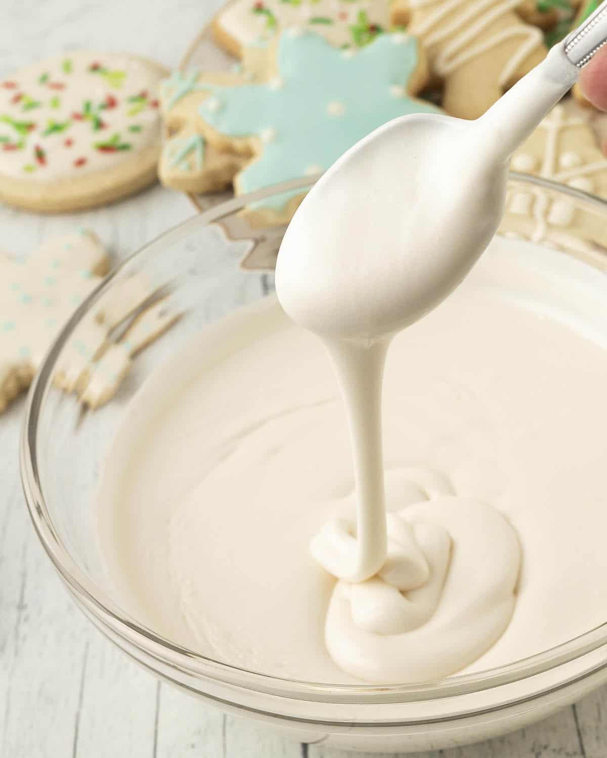 A bowl of eggless royal icing on a table, a spoon of icing is being held over the bowl, cookies sit behind the bowl.