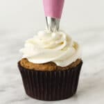 Cream cheese frosting being piped onto a carrot cupcake with a pink coloured piping bag.