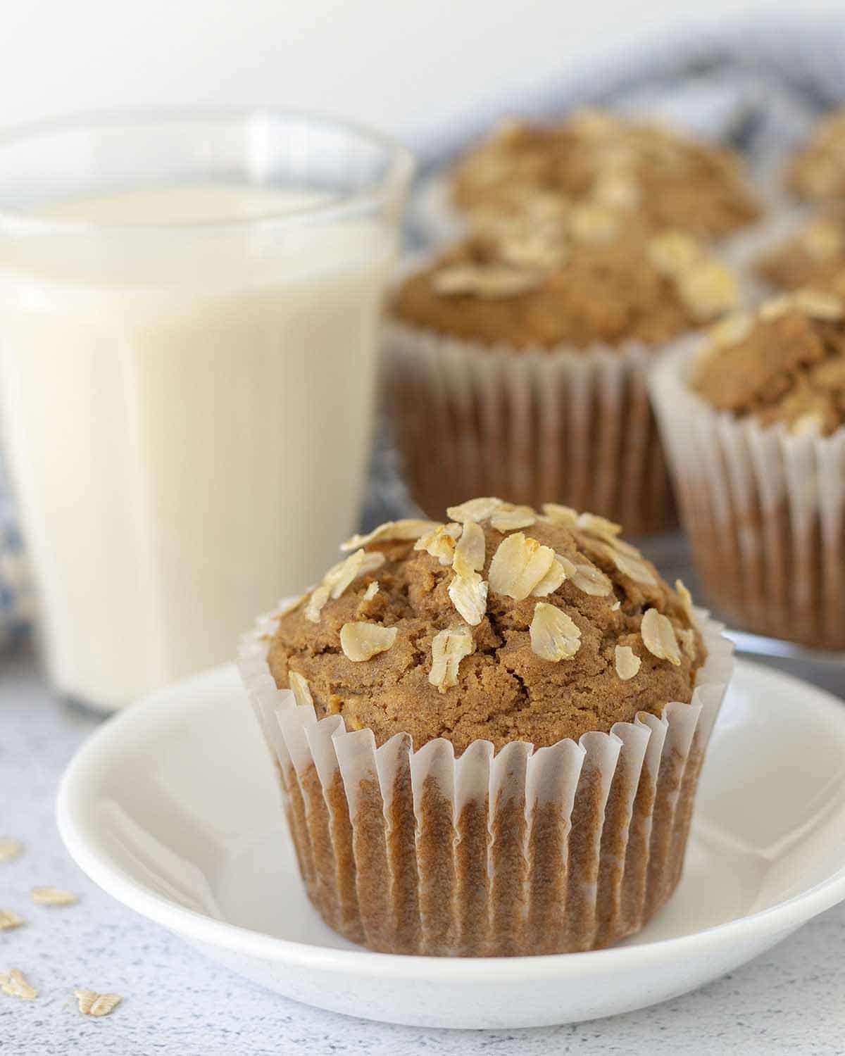 Image shows an oatmeal muffin on a white plate, more muffins sit a cooling rack in the background.