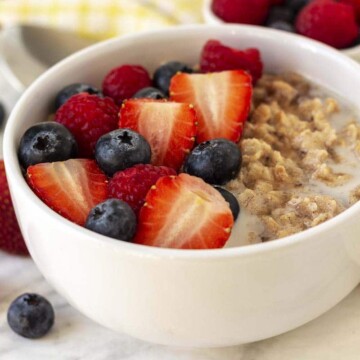 A bowl of flaxseed porridge on a white table, fresh berries top of the oatmeal and a few berries are also on the table.