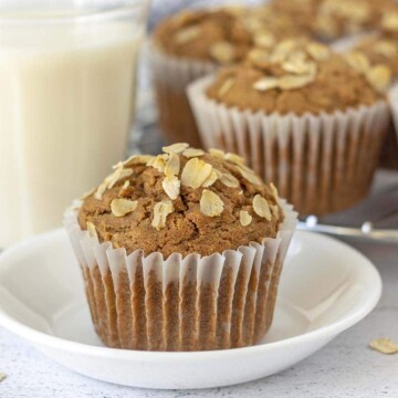 An oatmeal muffin sitting on a small white plate, more muffins and a glass of almond milk sit behind the plate.