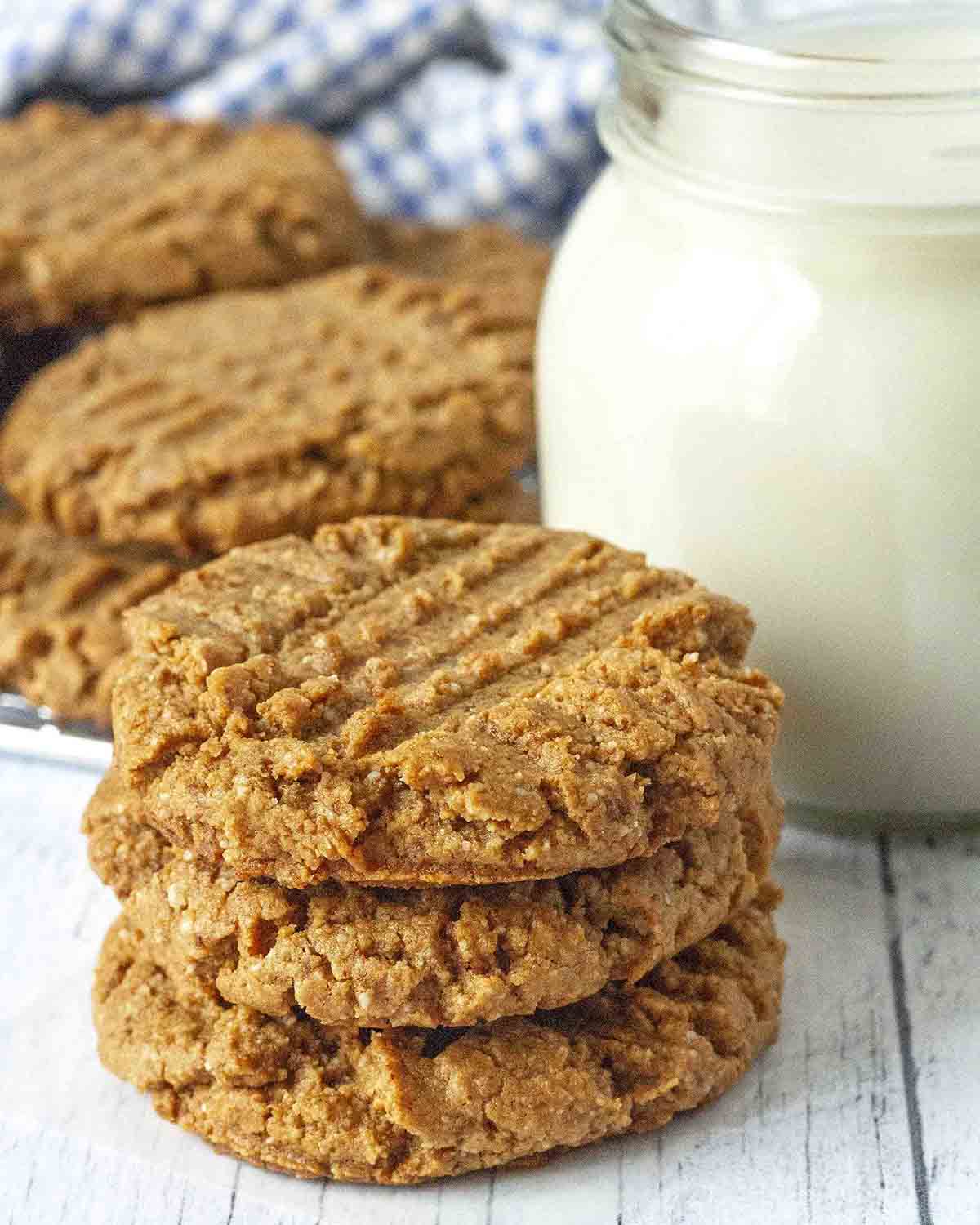 A stack of three gluten free peanut butter cookies on a white table, a glass of almond milk sits behind the stack.