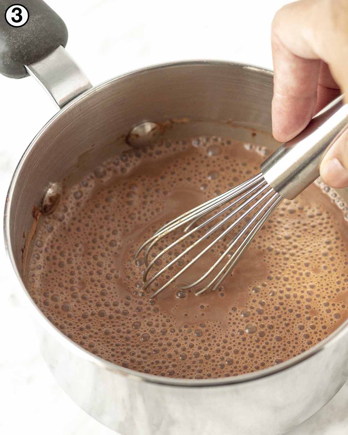 A hand using a metal whisk to mix hot chocolate in a small pot.
