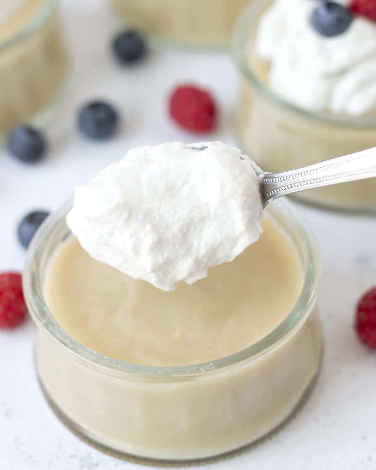 An image showing a spoon putting dairy-free whipped cream on top of a bowl of eggless vanilla pudding.