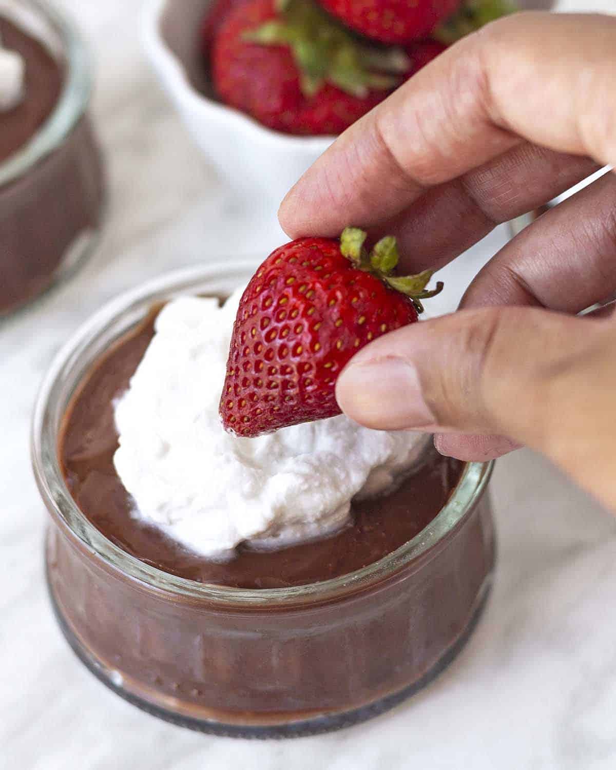 A hand putting a fresh strawberry on top of a small bowl of eggless chocolate pudding.