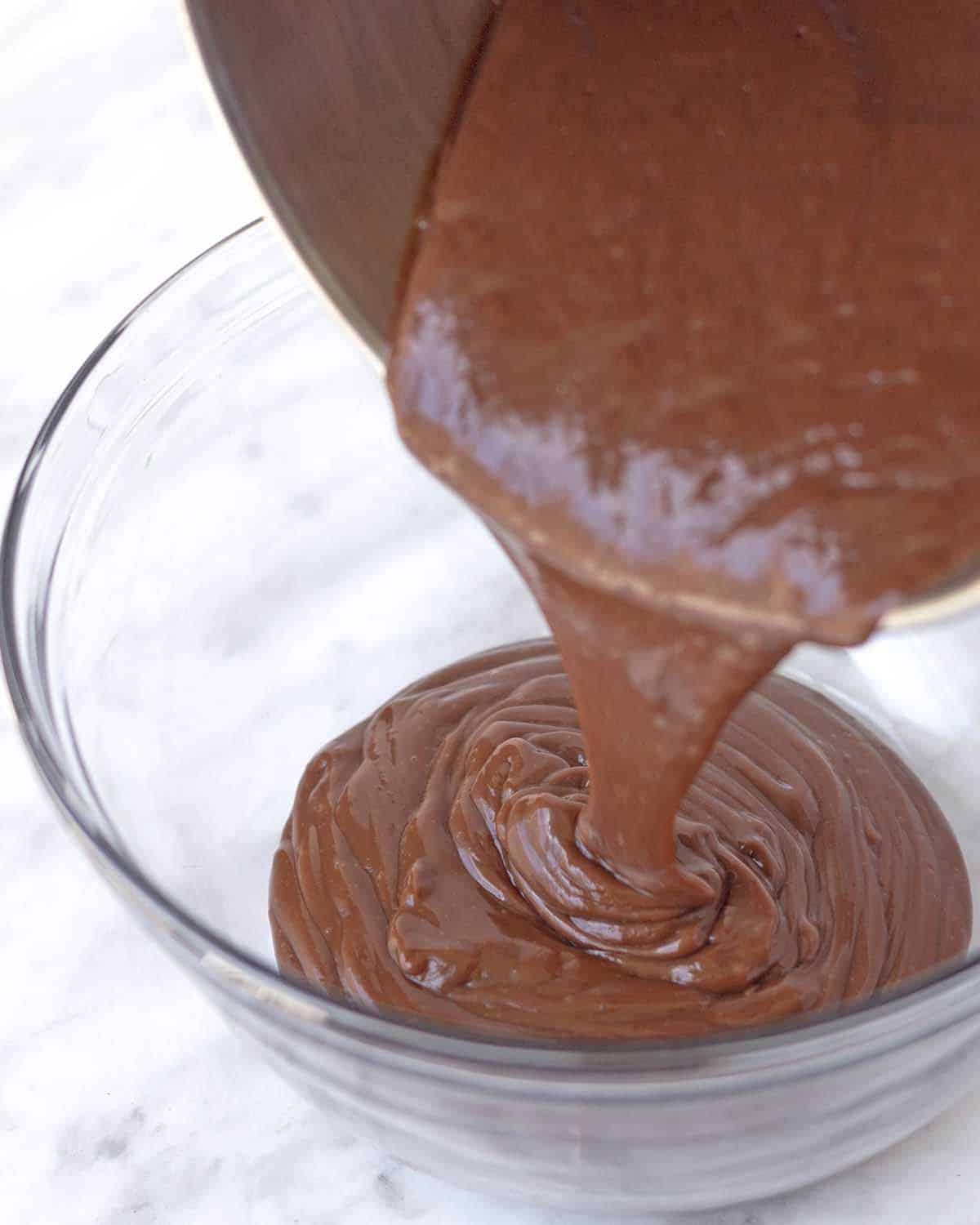 Homemade vegan gluten-free chocolate pudding being poured from a pot into a glass bowl.