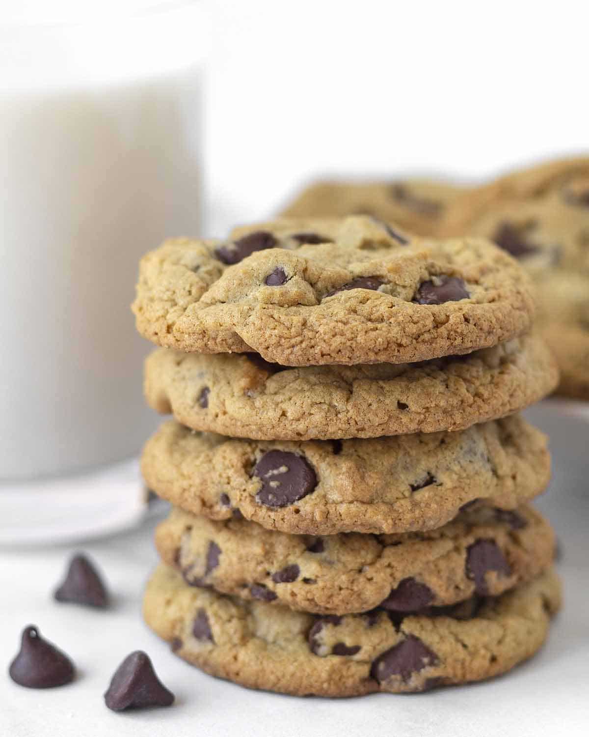 A stack of five oat flour cookies, a glass of milk and more cookies can be seen behind the stack.