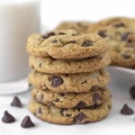 A stack of five chocolate chip cookies made with oat flour on a white table, a glass of milk and more cookies sit behind.