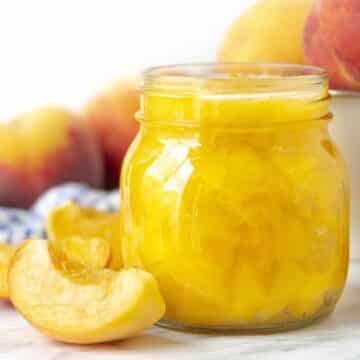 A glass jar of homemade peach sauce sitting on a table surrounded by fresh peaches.