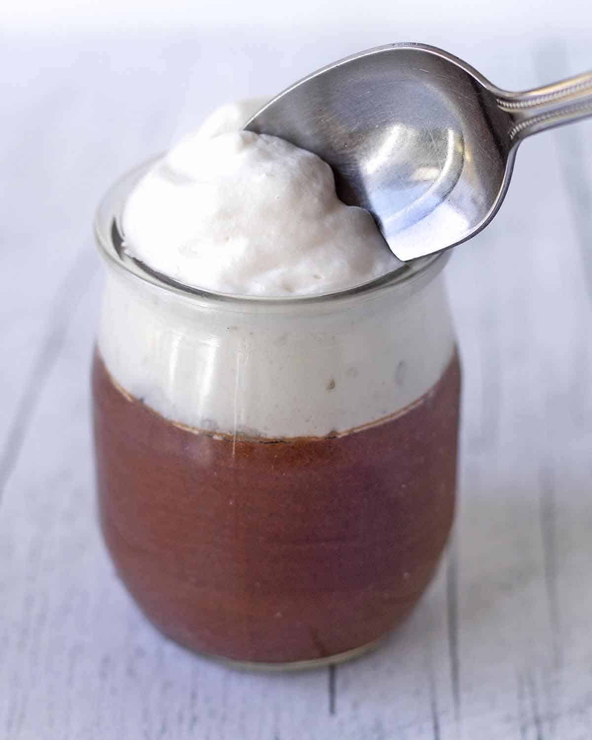 A small jar filled with chocolate mousse topped with vegan whipped cream, a spoon is taking out a spoonful of the dessert.