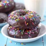 A close-up image showing two baked chocolate donuts with sprinkles sitting on a small white plate.