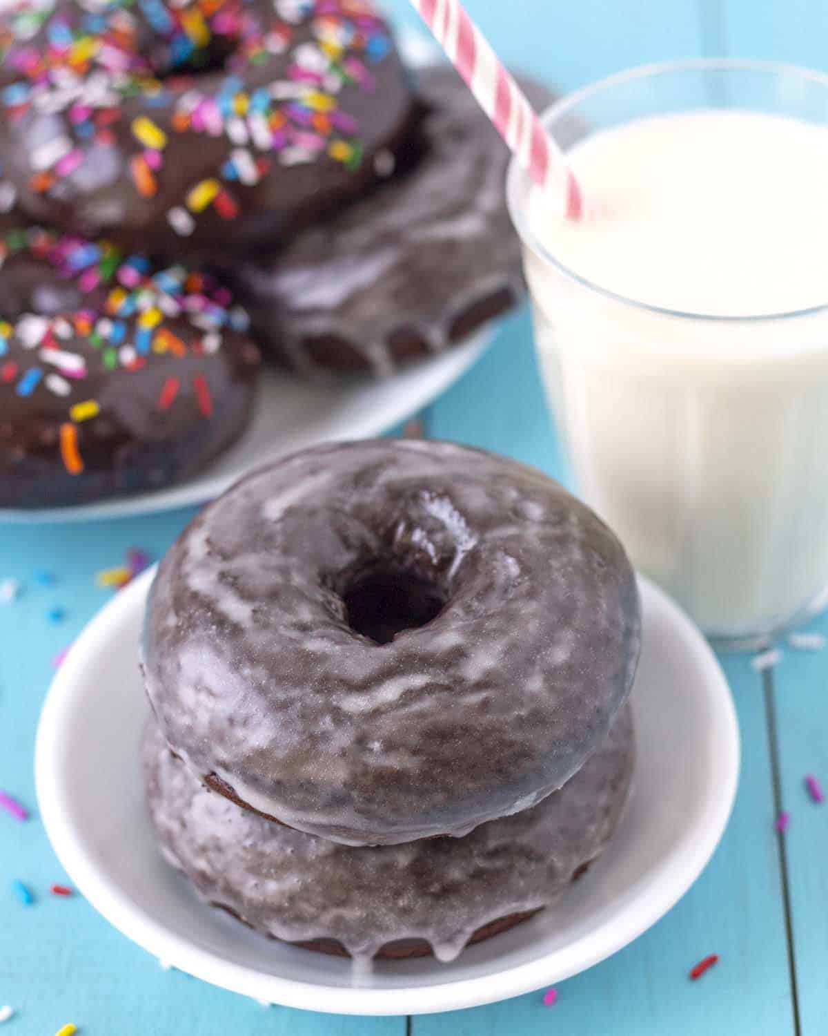 Two vanilla sugar glaze dipped vegan chocolate donuts with sprinkles sitting on a small white plate.