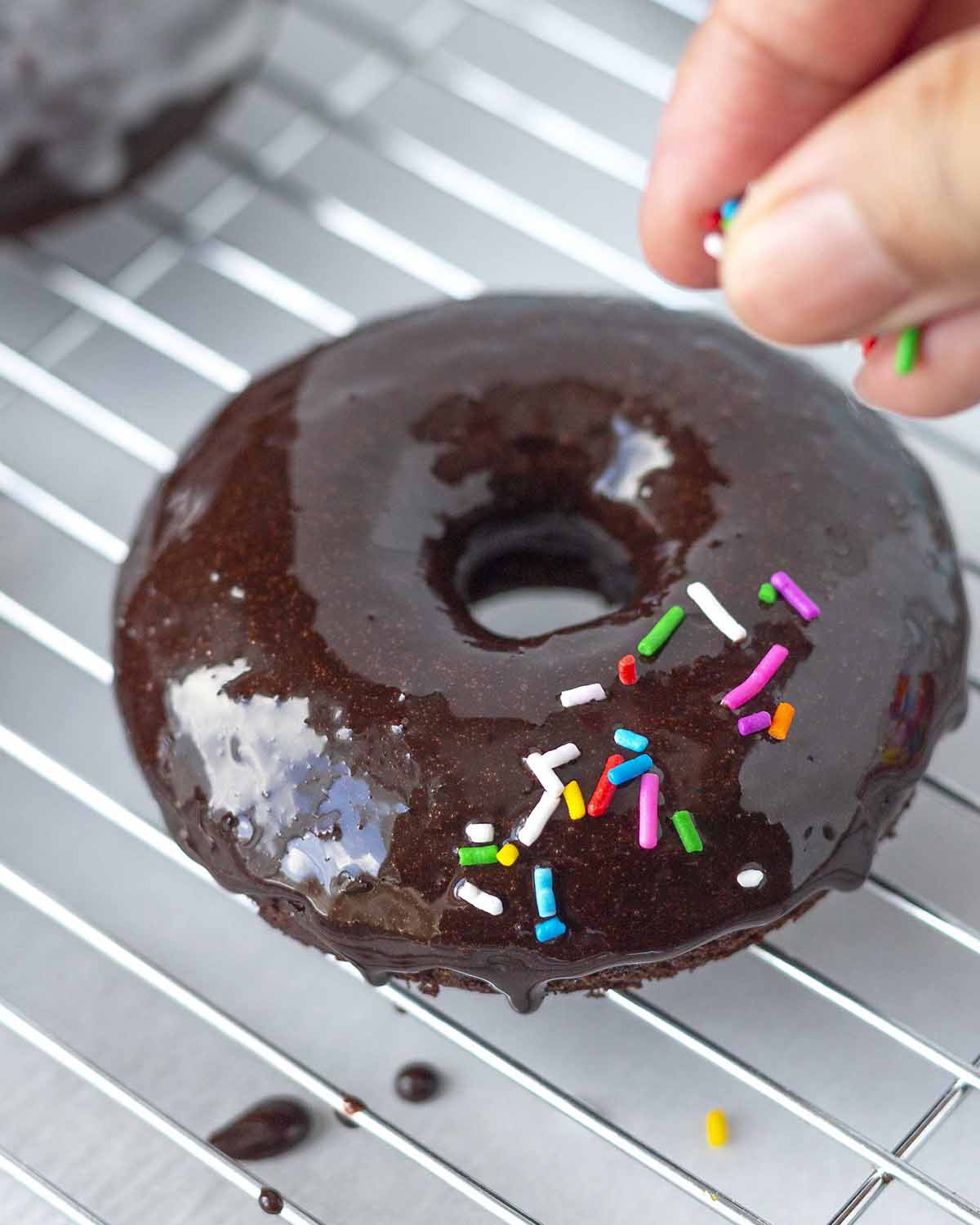 A hand adding sprinkles to a freshly dipped chocolate donut.