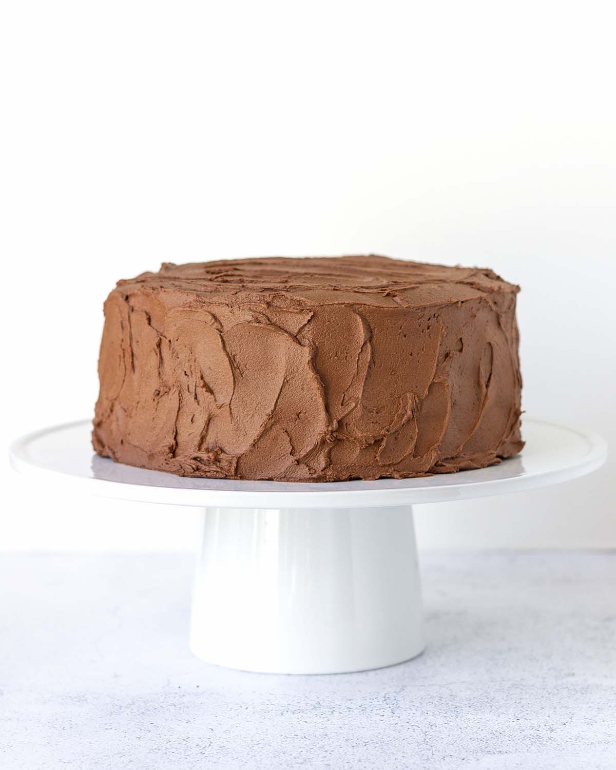 A gluten-free vegan chocolate cake frosted with chocolate buttercream sitting on a white ceramic cake stand.
