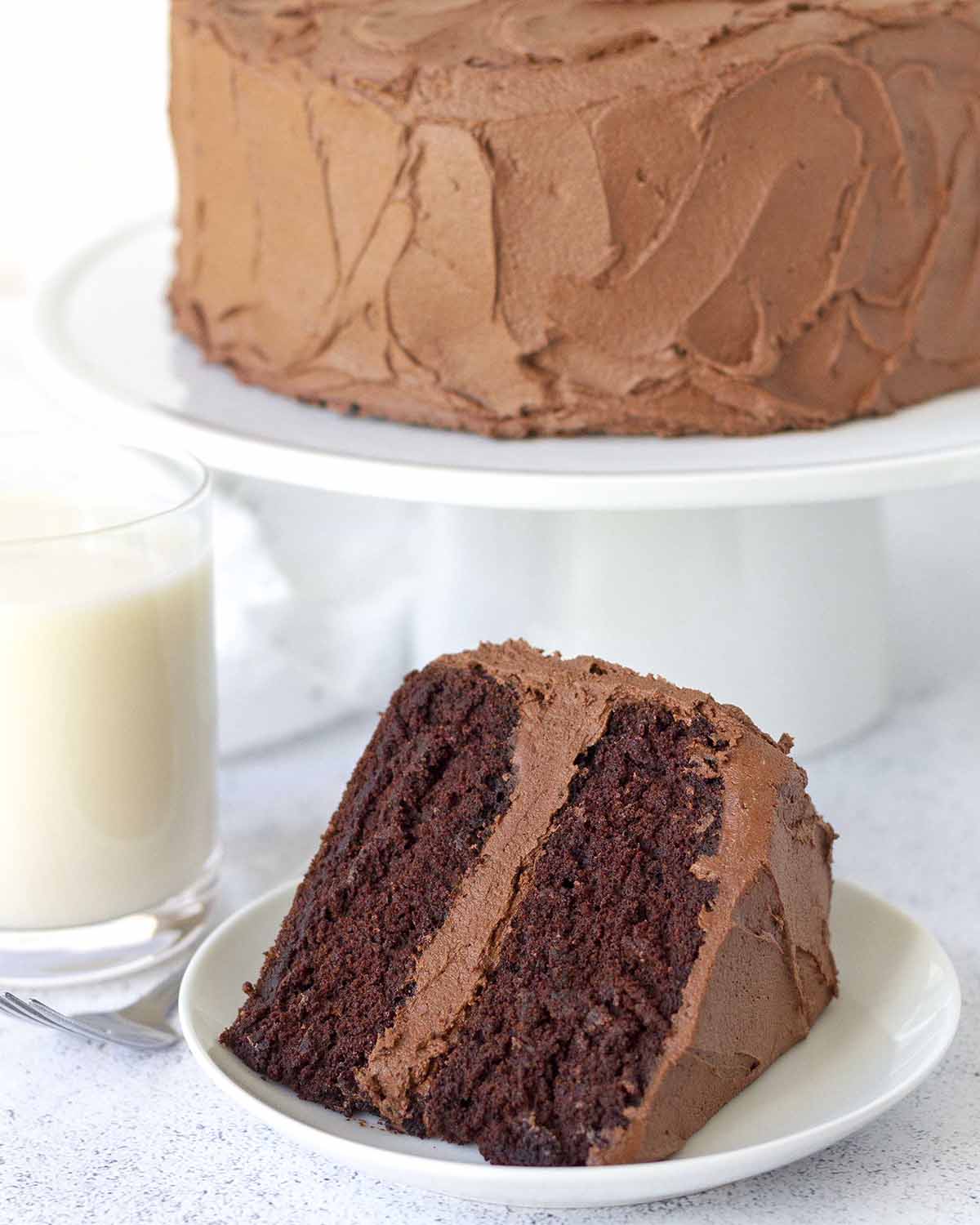 A slice of frosted chocolate layer cake on a plate, the rest of the cake sits behind the plate on a white cake stand.