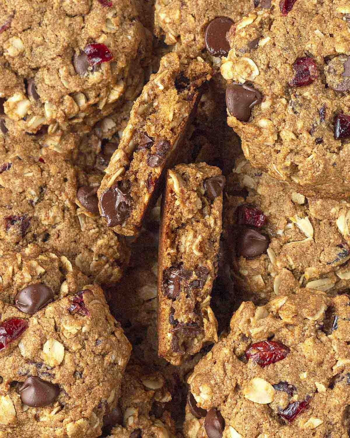 A plate full of vegan oatmeal cranberry cookies with chocolate chips, one cookie has been broken to show the inner texture.