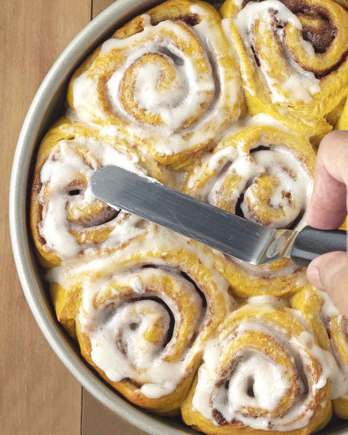 A hand frosting a pan of freshly baked vegan pumpkin cinnamon rolls with a small offset spatula.