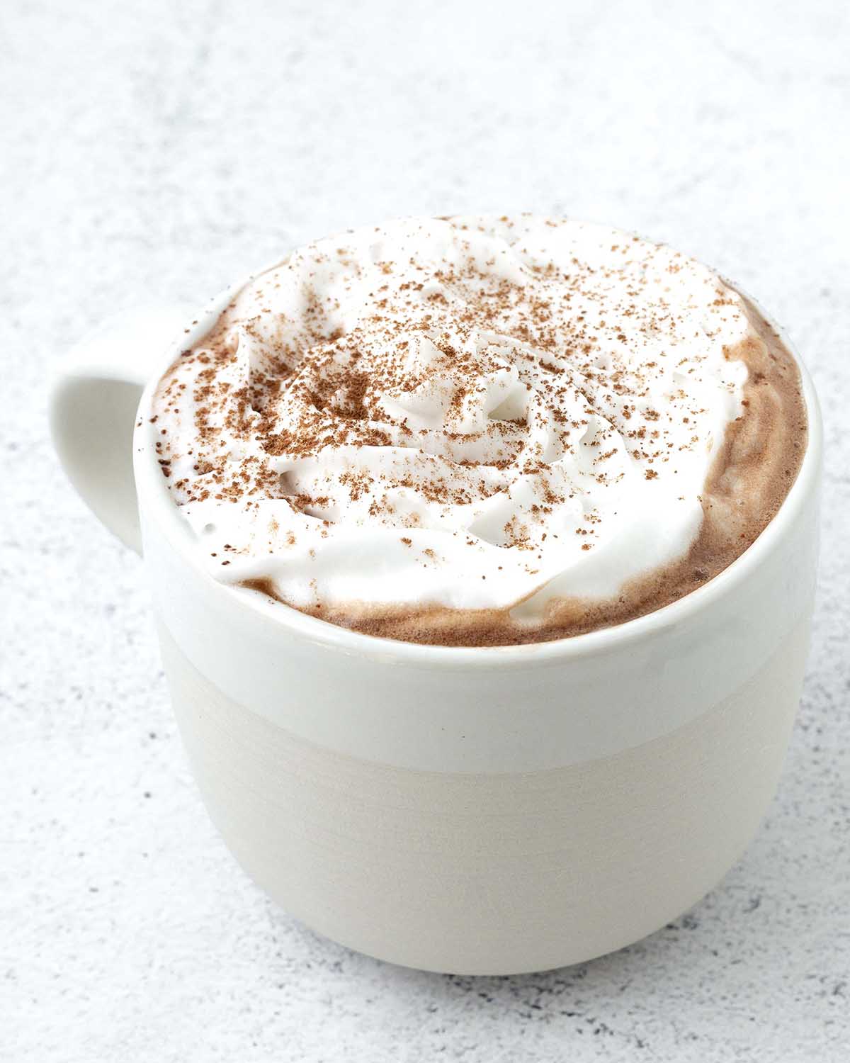 A mug of non-dairy hot chocolate in a mug, it is garnished with vegan whipped cream and a dusting of cocoa powder.