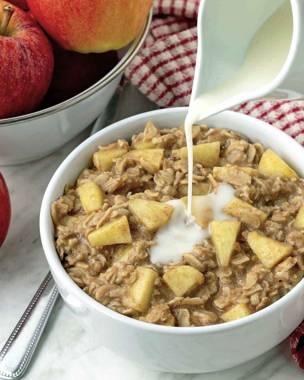 Milk being poured from a white ceramic jar onto a bowl of apple cinnamon oatmeal.