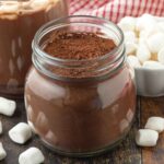 A glass jar of homemade hot cocoa mix sitting on a dark wood table, mini marshmallows are also on the table.