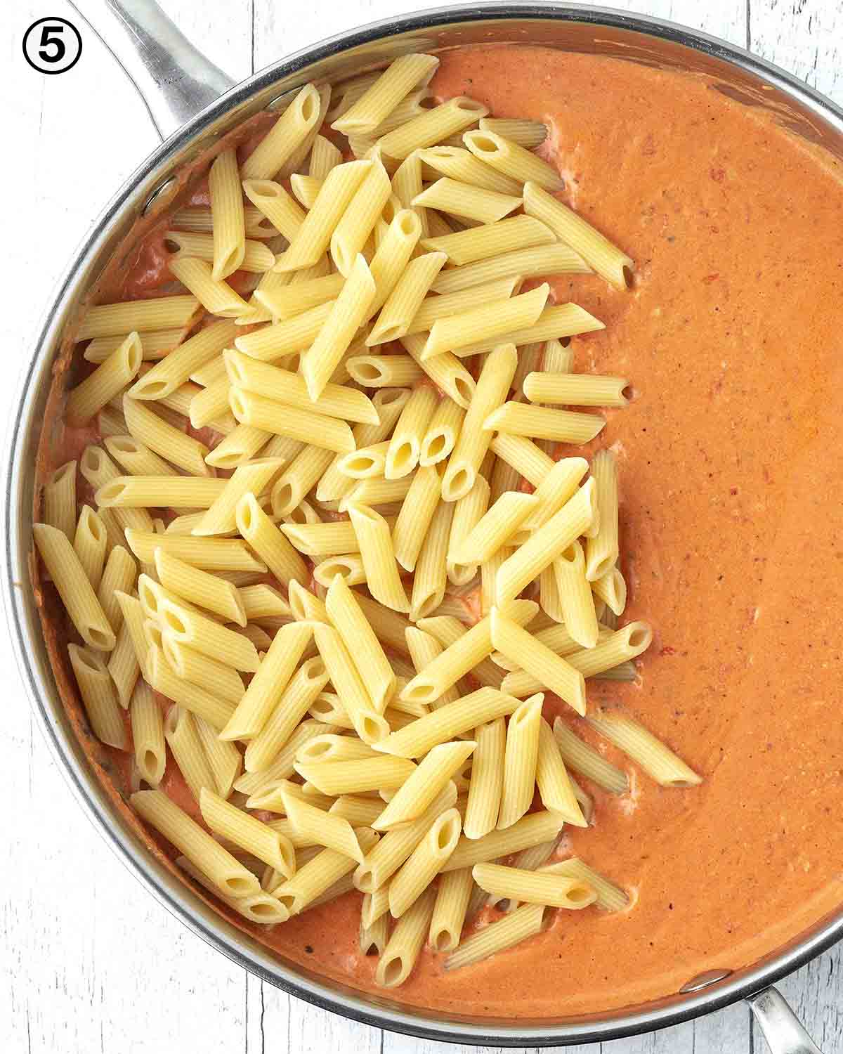 An overhead shot showing pasta sauce in a pan with cooked penne pasta on top waiting to be stirred in.