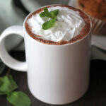 A mug of peppermint hot cocoa garnished with whipped cream and fresh mint leaves.