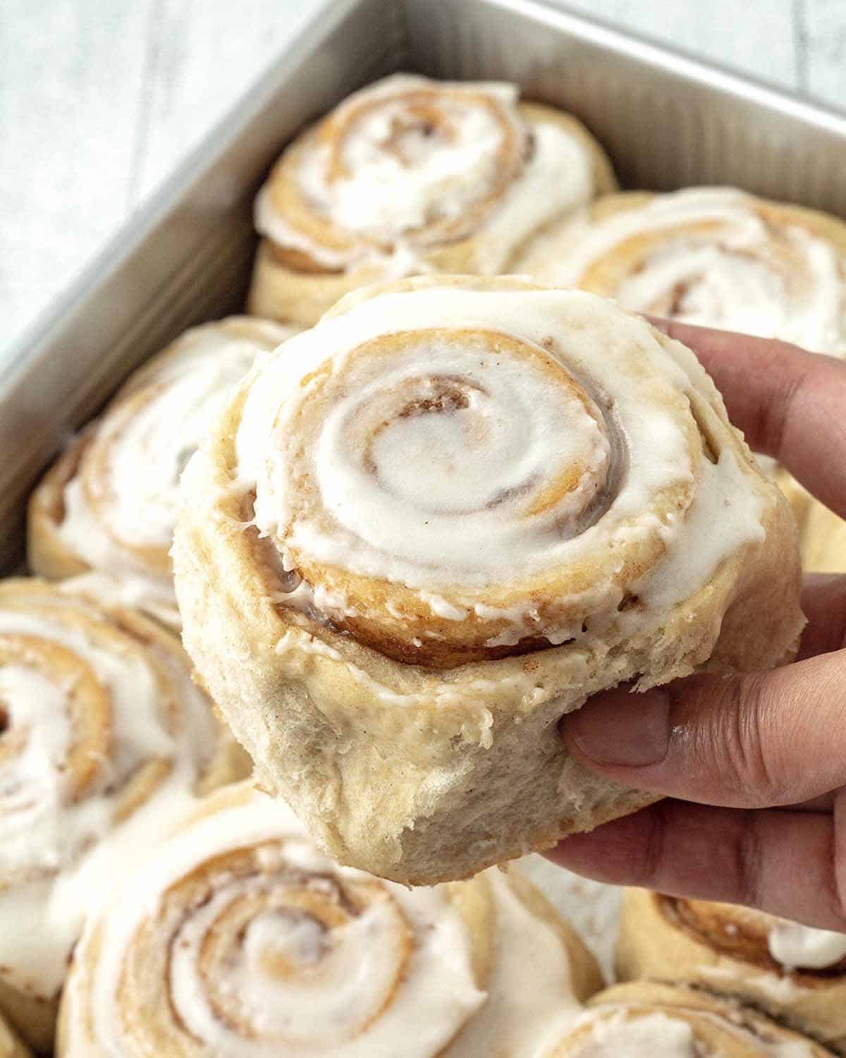 A hand taking a freshly baked cinnamon roll out of the baking pan.