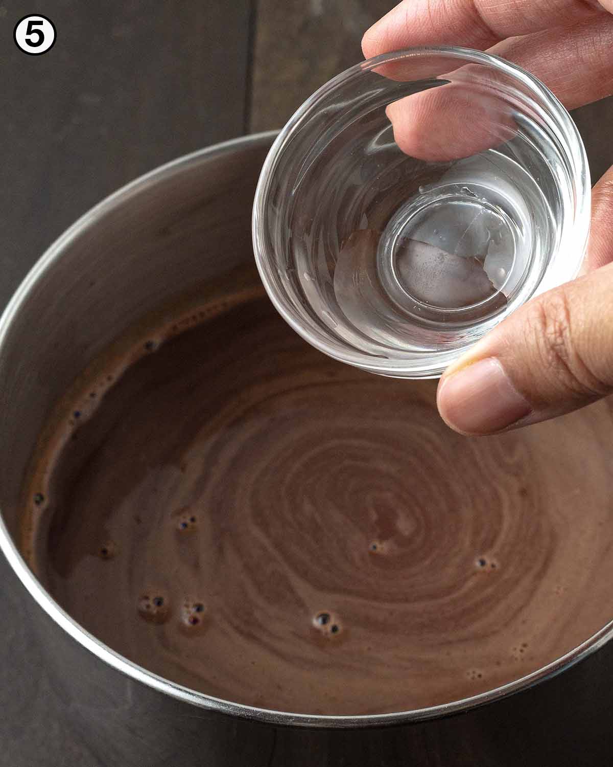 A hand pouring peppermint extract from a small glass bowl into a pot of hot chocolate.