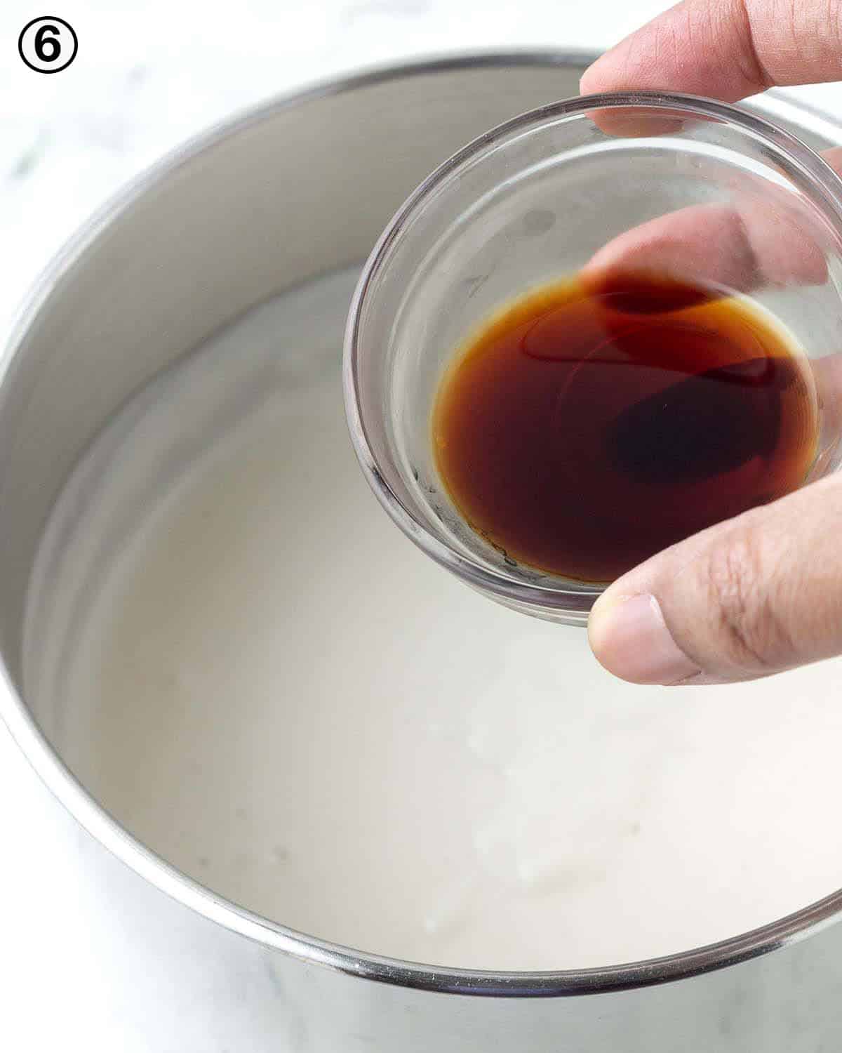 A hand pouring vanilla extract from a small glass bowl into a pot of dairy-free vanilla custard.