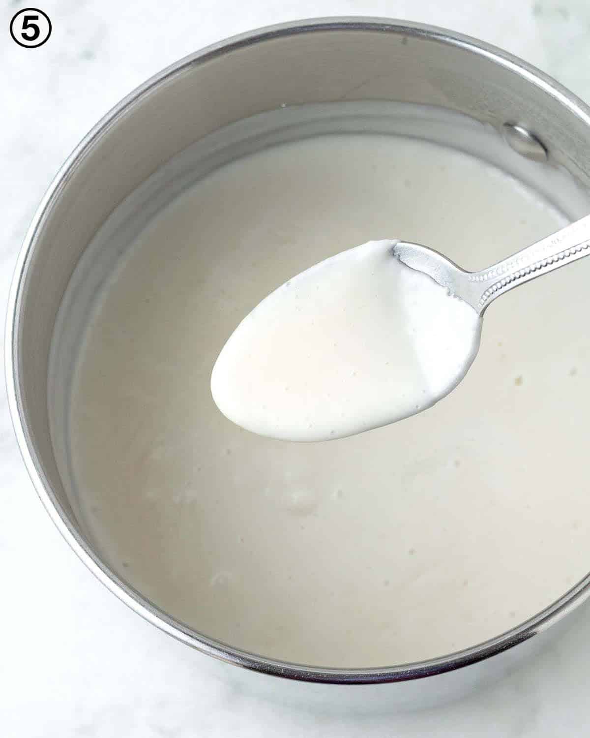A spoon being held over a pot of cooked vanilla custard, the custard has thickened and is clinging to the spoon.