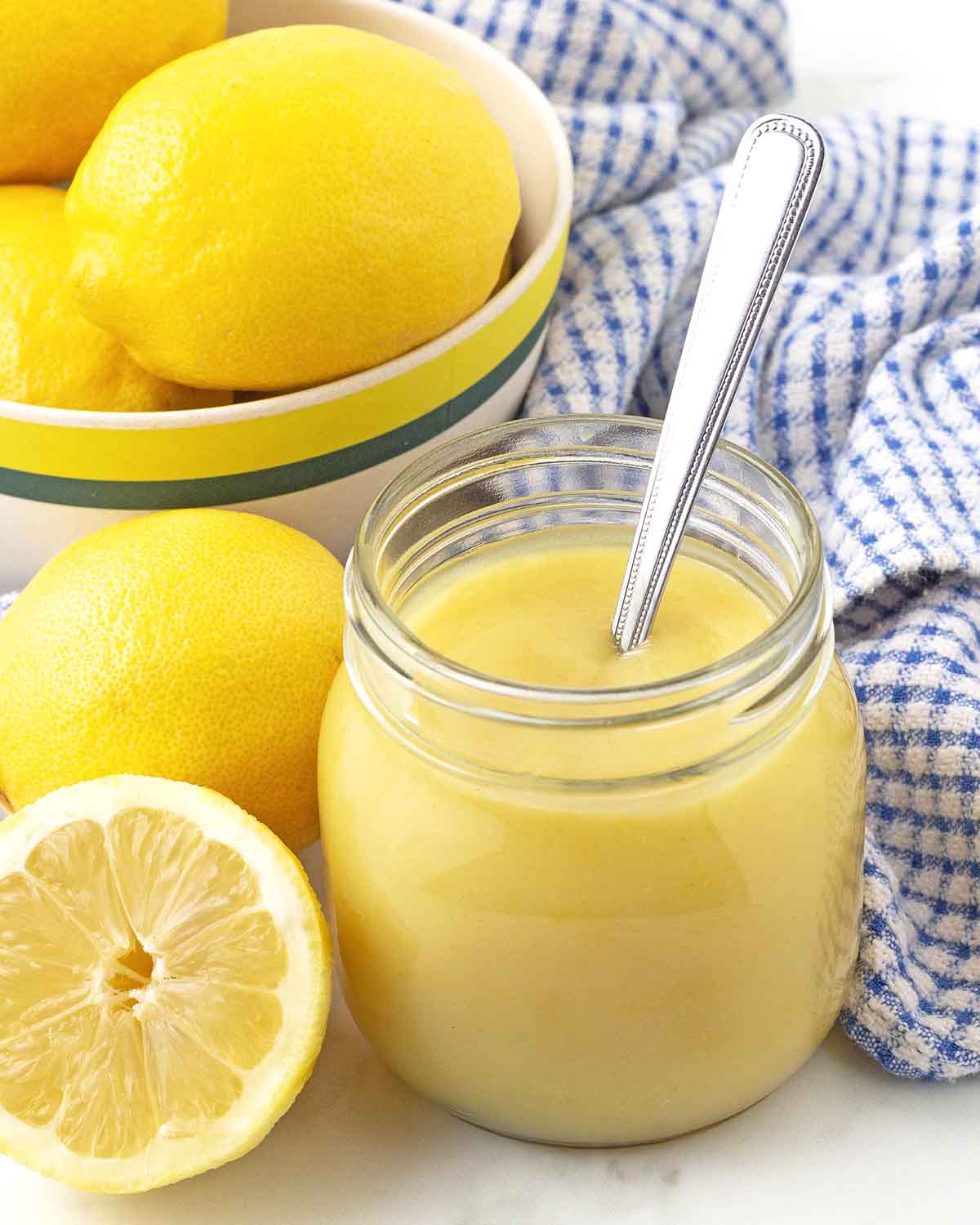 A jar of lemon curd with a spoon in it, fresh lemons sit to the left and a bowl of lemons sits behind the jar.