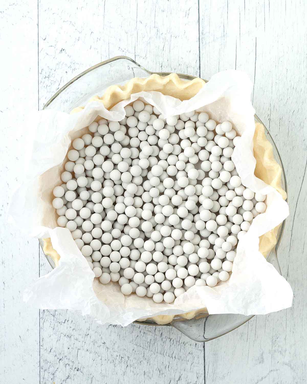 An overhead shot of a raw pie crust in a glass pie dish lined with parchment paper and filled with ceramic pie weights.