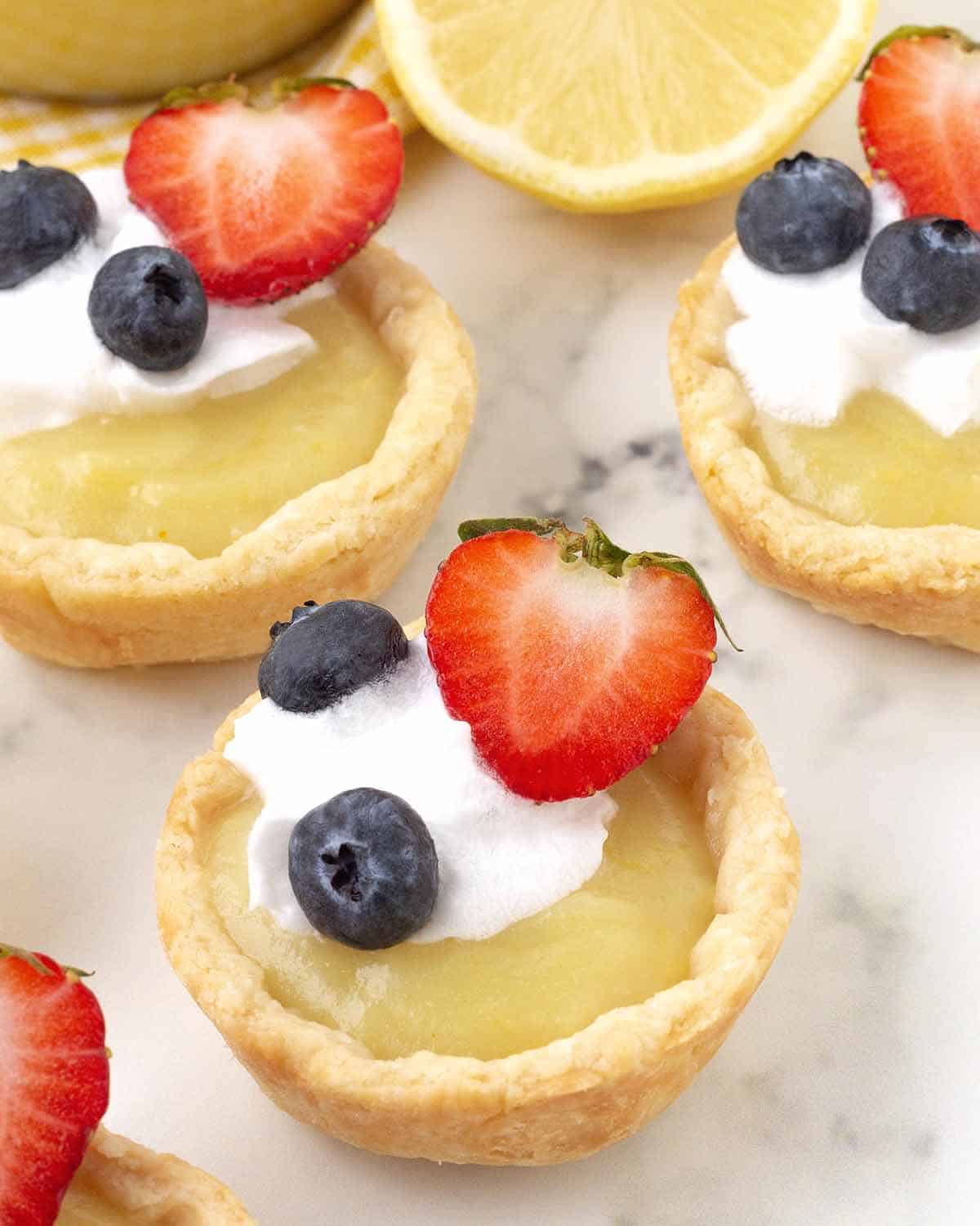 Mini vegan lemon curd tarts on a table, tarts are garnished with coconut whip and fresh berries.