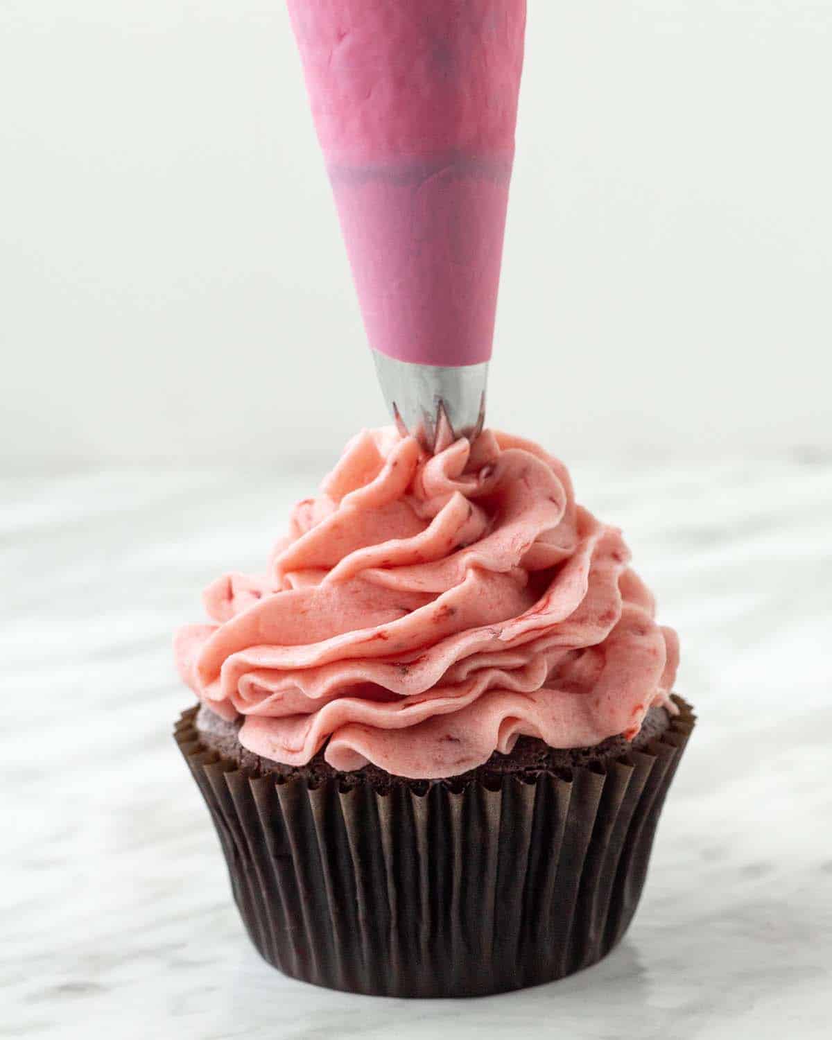 Homemade strawberry buttercream being piped onto a chocolate cupcake.
