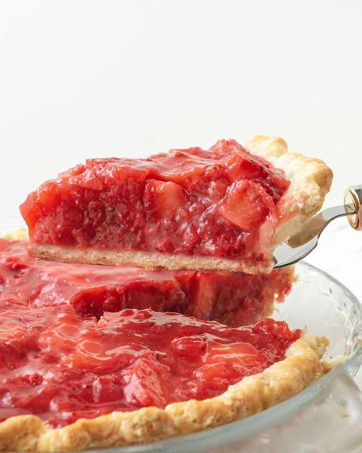 A slice of strawberry pie being lifted out of a pie plate and away from the rest of the pie with a pie server.