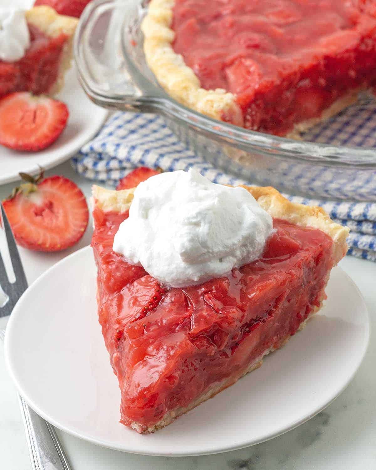 A slice of strawberry pie on a white plate, the pie is topped with whipped cream.
