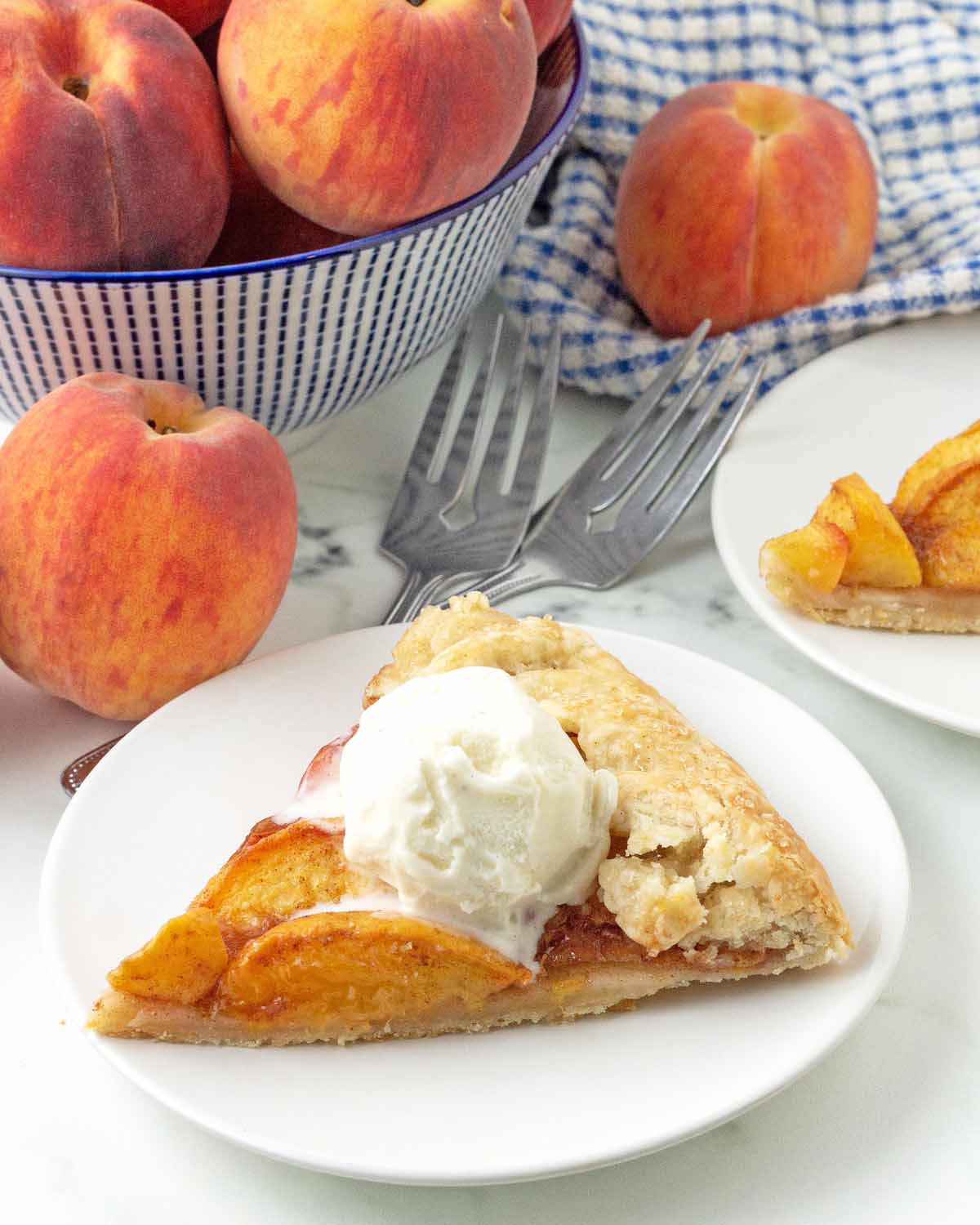 A slice of a peach galette on a plate is topped with ice cream, and fresh peaches sit in the background.